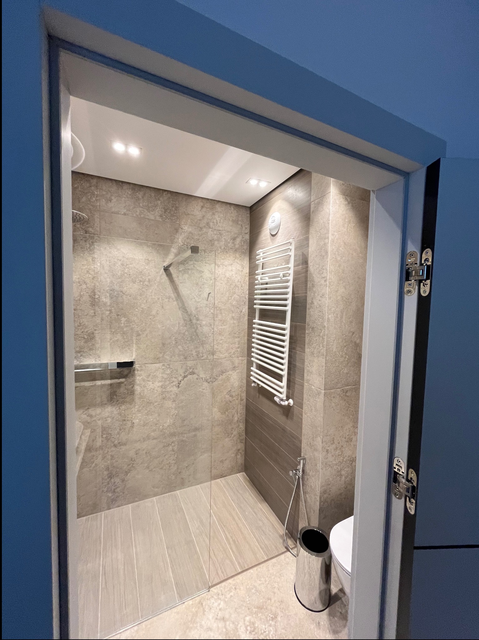 Nowoczesna łazienka z kabiną prysznicową typu walk-in, grzejnikiem drabinkowym i bidetką. Uwagę zwraca minimalistyczny design i stonowana kolorystyka płytek.