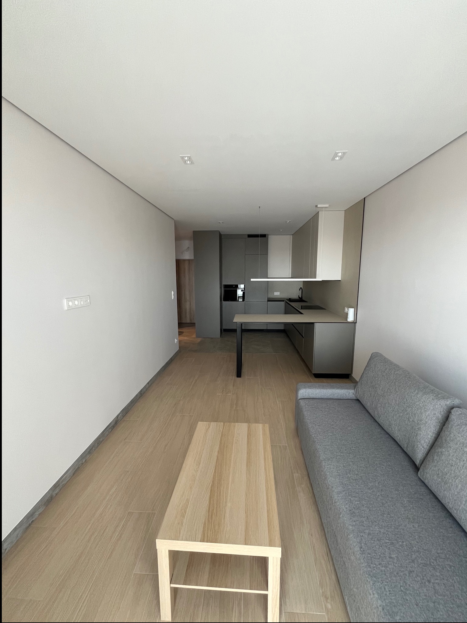 Nowoczesne wnętrze: szara kuchnia z wyspą, drewniany stół kawowy i szara sofa. Minimalistyczny design z drewnianą podłogą i białymi ścianami.