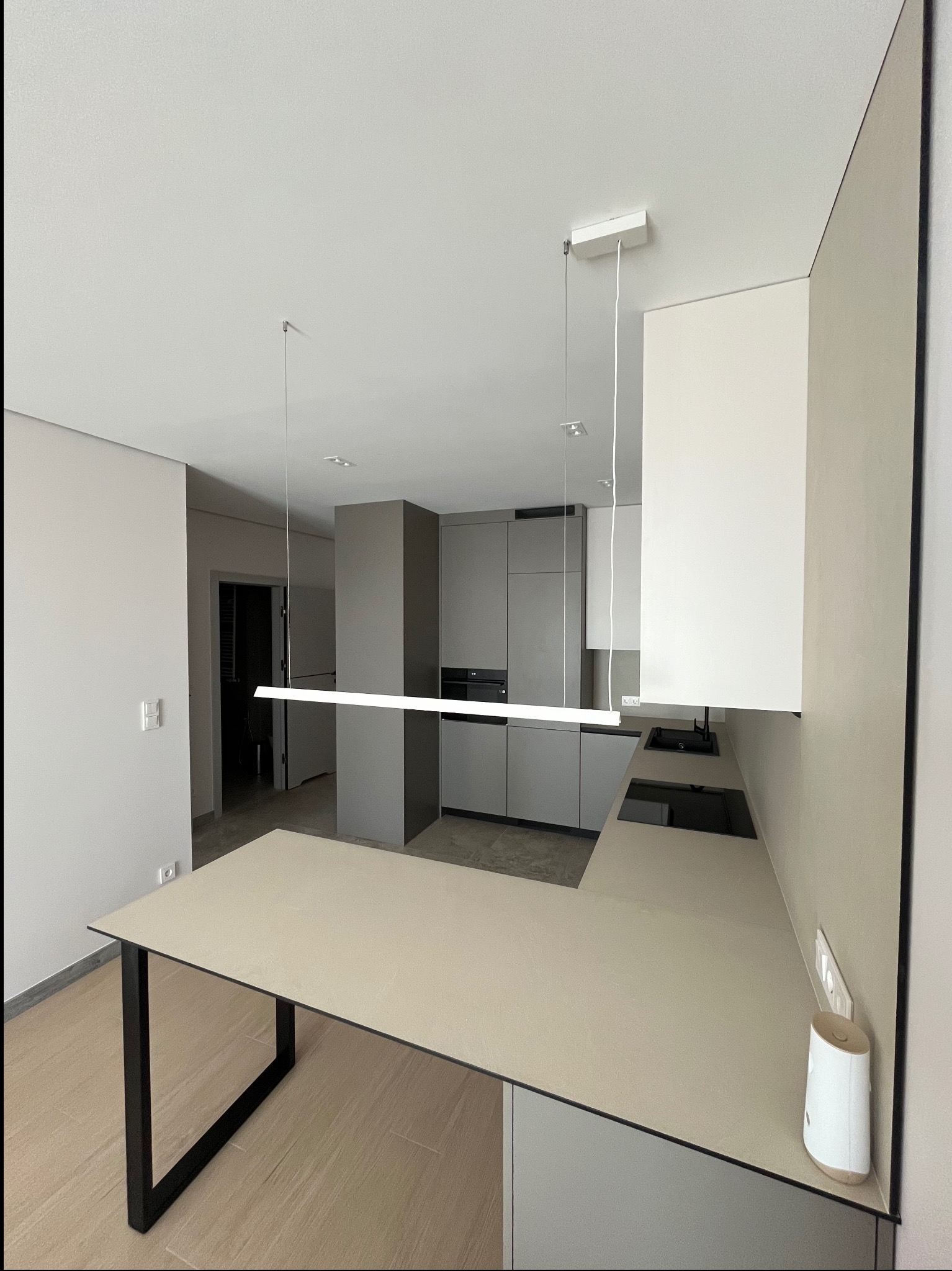 Nowoczesna, jasna kuchnia w odcieniach szarości i beżu z minimalistycznym oświetleniem LED. Widoczne szafki, blat i stół z czarnymi nogami. W tle otwarte drzwi.