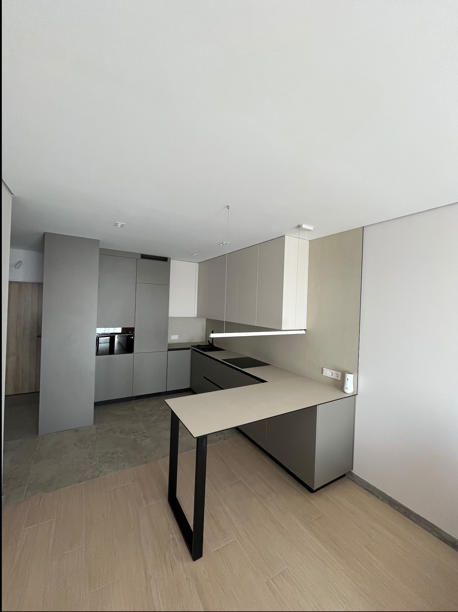 Nowoczesna kuchnia w odcieniach szarości, z wyspą i minimalistycznym oświetleniem. Widoczne płytki podłogowe imitujące drewno i kamień. Ujęcie z perspektywy.