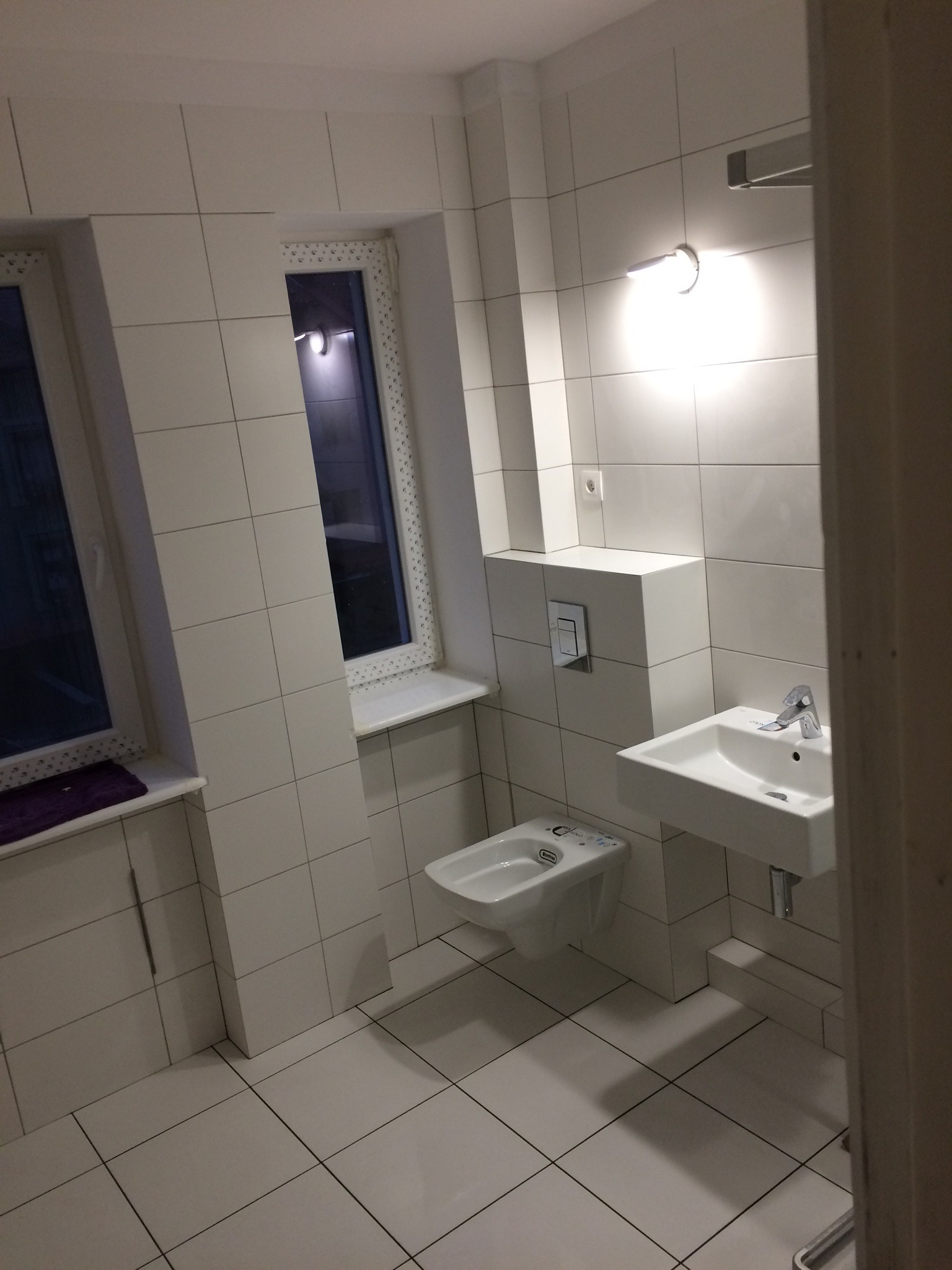 Wykończona łazienka w bieli z kwadratowymi płytkami, wiszącą toaletą i umywalką, oświetlona lampą nad lustrem, z widocznymi oknami po lewej stronie.