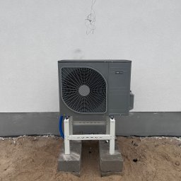 KGK INSTALACJE KAJETAN MIKOŁAJCZAK - Szara pompa ciepła Nibe zamontowana na białym stelażu na betonowych bloczkach, na tle białej ściany budynku. Widoczne niepodłączone przewody.
