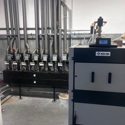 KGK INSTALACJE KAJETAN MIKOŁAJCZAK - Nowoczesna kotłownia z szarym kotłem Metal-Fach i rzędem izolowanych rur. Widoczne liczniki i zawory. Minimalistyczne wnętrze z betonową podłogą i białymi ścianami.