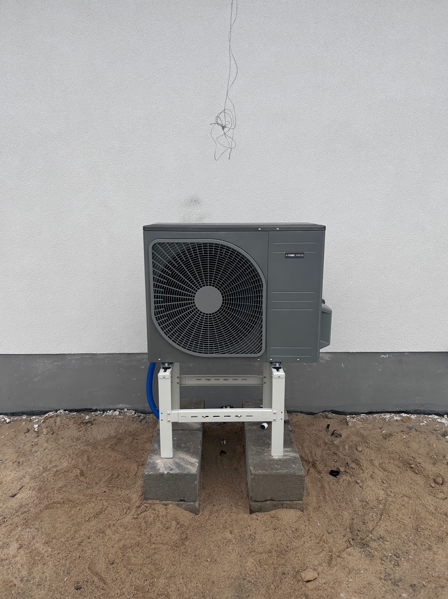 Szara pompa ciepła Nibe zamontowana na białym stelażu na betonowych bloczkach, na tle białej ściany budynku. Widoczne niepodłączone przewody.