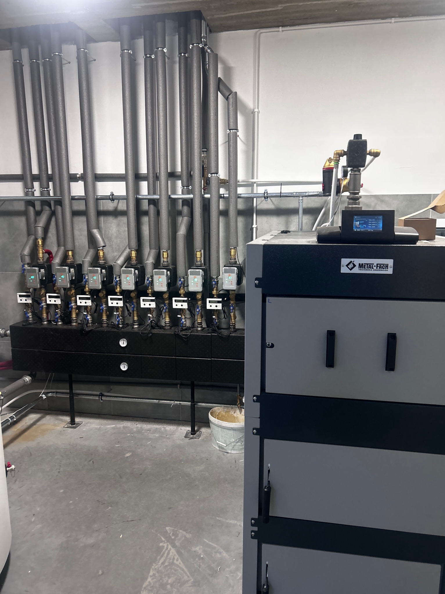Nowoczesna kotłownia z szarym kotłem Metal-Fach i rzędem izolowanych rur. Widoczne liczniki i zawory. Minimalistyczne wnętrze z betonową podłogą i białymi ścianami.