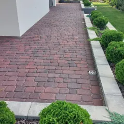 Czerwono-brązowa kostka brukowa tworząca ścieżkę wzdłuż białego budynku, obrzeża z szarego betonu, ozdobne krzewy i trawnik po prawej stronie, pergola w tle.