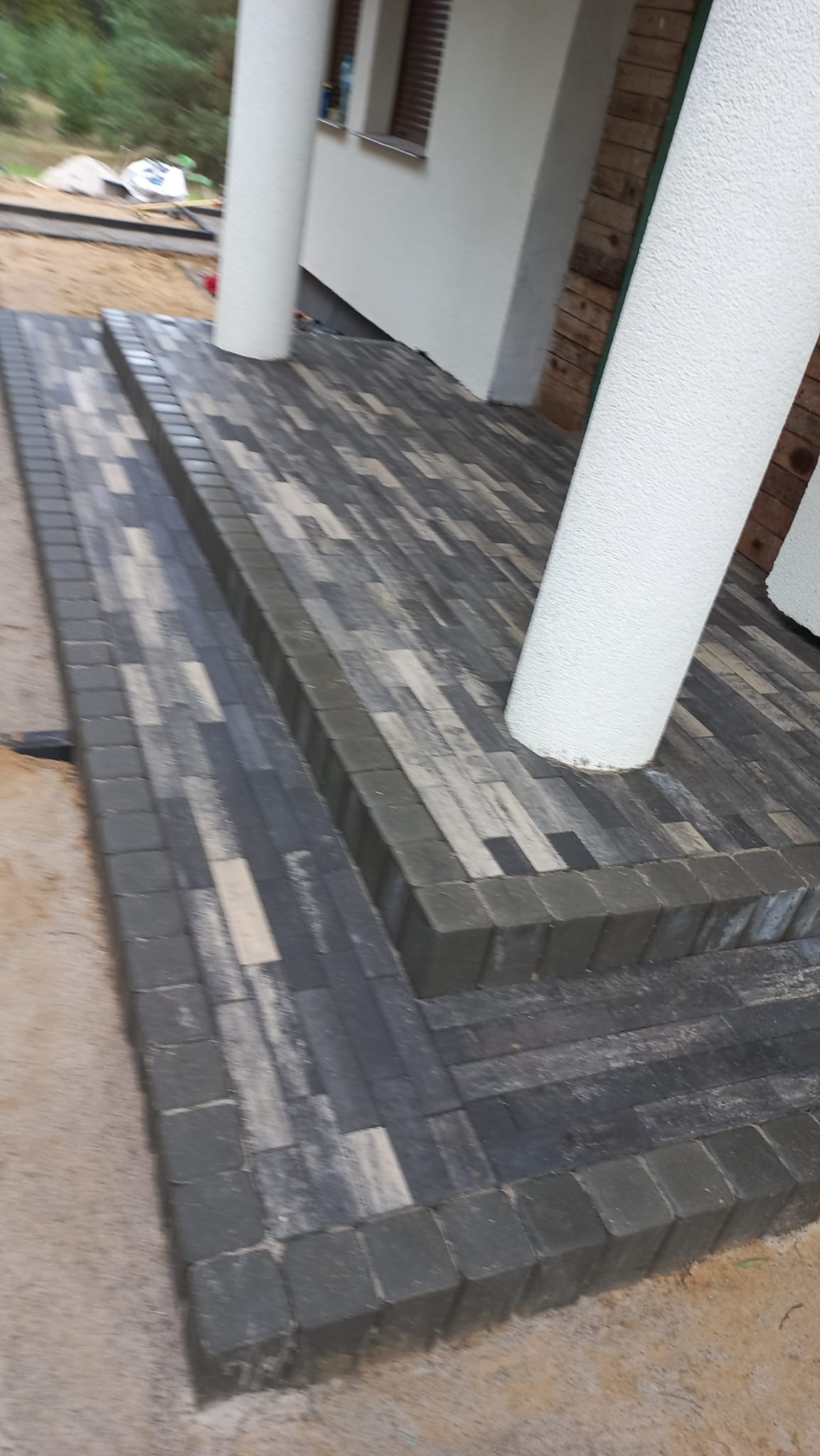 Schody wejściowe wyłożone kostką brukową w odcieniach szarości i bieli, z widocznymi kolumnami podtrzymującymi zadaszenie.