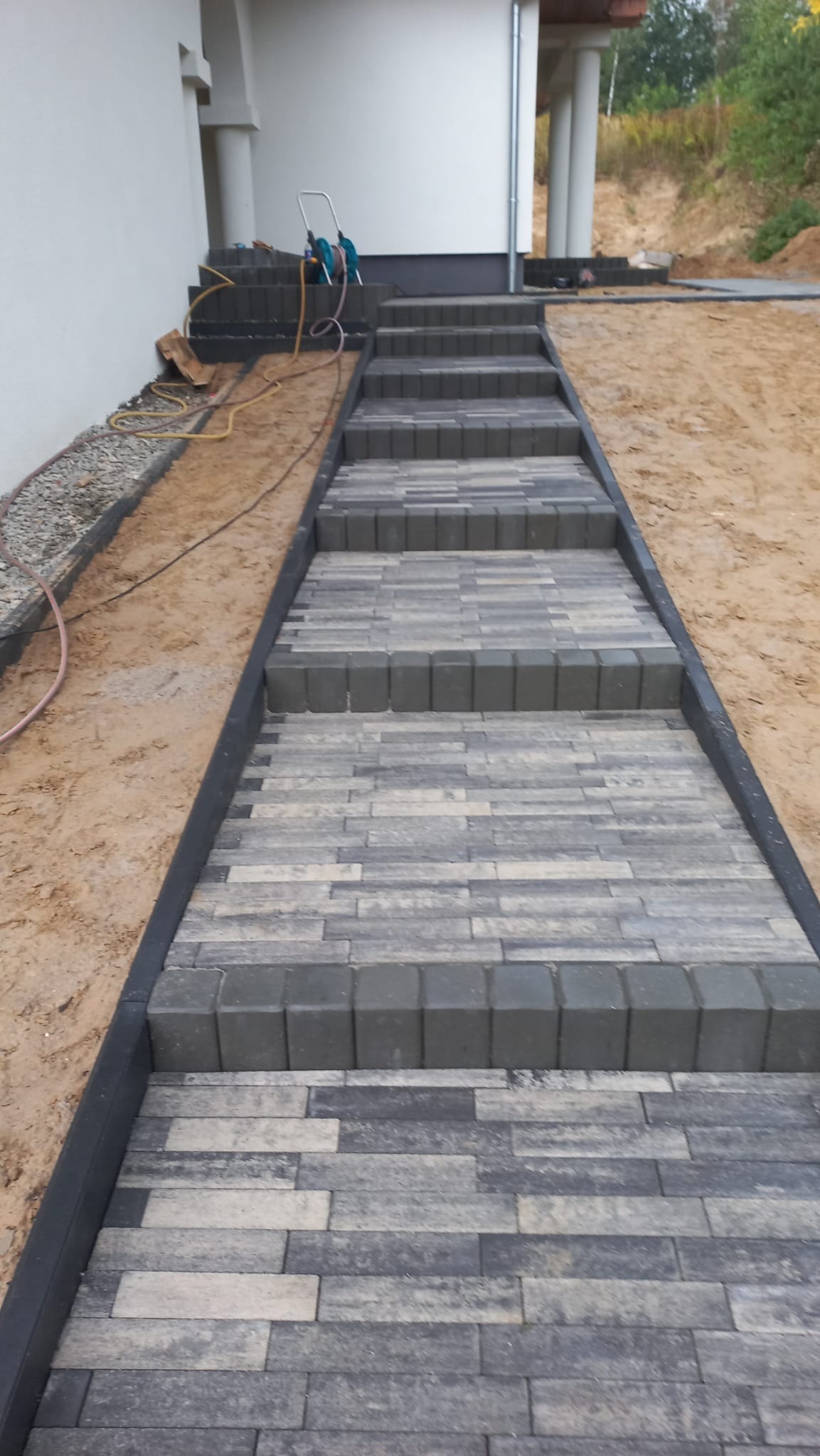 Schody wejściowe wyłożone szaro-beżową kostką brukową, z czarną obwódką, prowadzące do budynku, z widocznym sprzętem budowlanym na górze schodów.