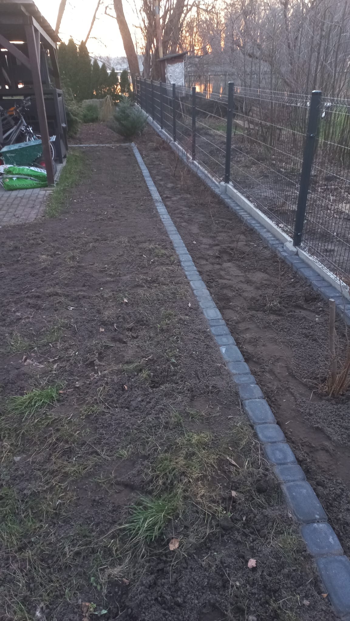 Przygotowany teren pod nasadzenia z obrzeżem z kostki brukowej i zamontowanym ogrodzeniem panelowym w tle, widok z perspektywy ziemi.
