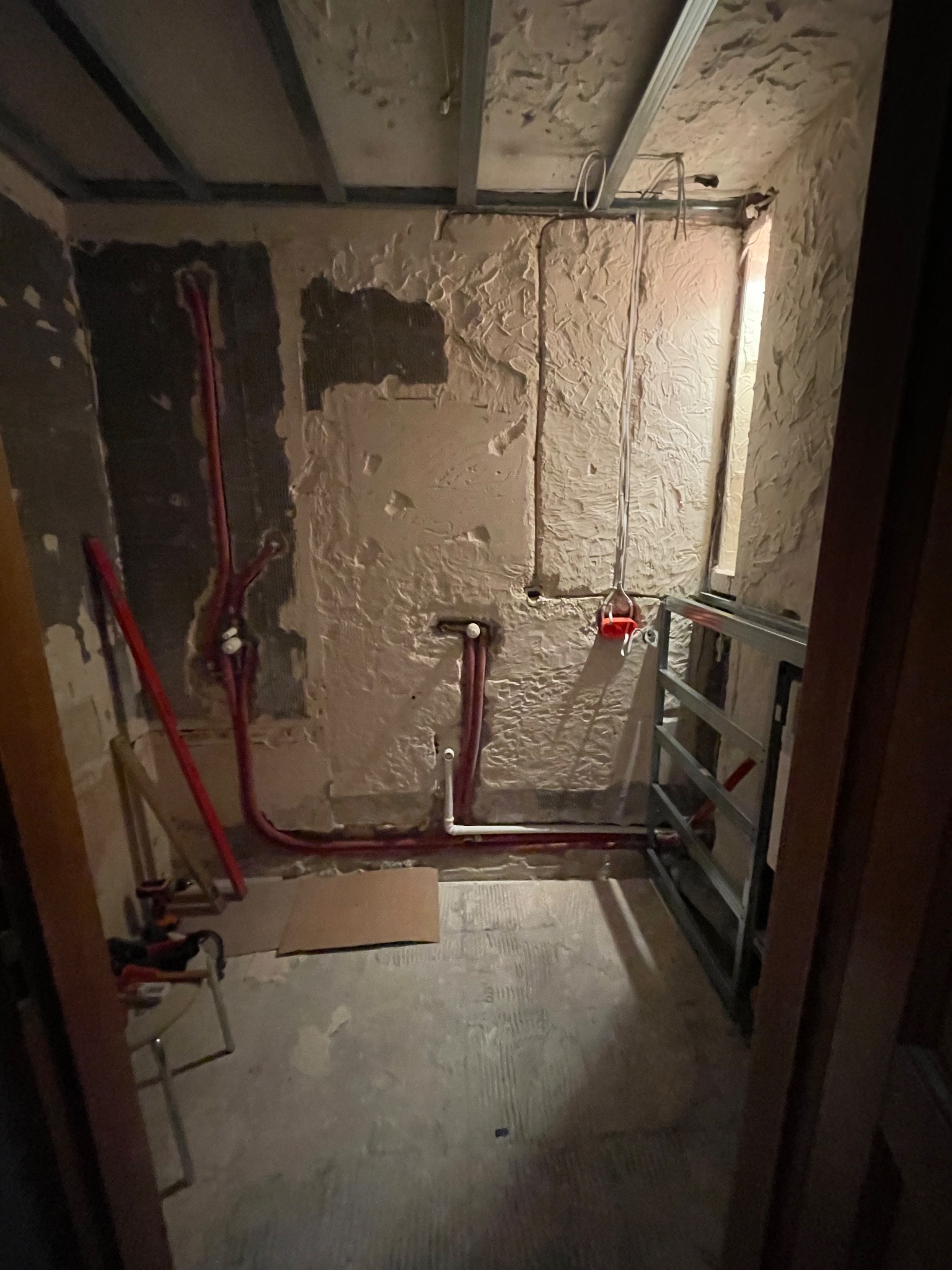 Wnętrze łazienki w trakcie prac: widoczne czerwone rury instalacji wodnej na ścianie z surowym tynkiem oraz metalowa konstrukcja pod zabudowę.