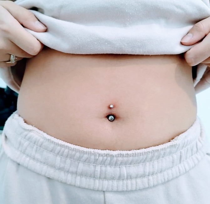 Zbliżenie na brzuch z dwoma kolczykami w pępku, jeden powyżej i jeden w samym pępku. Osoba w jasnej bluzie i spodniach dresowych.