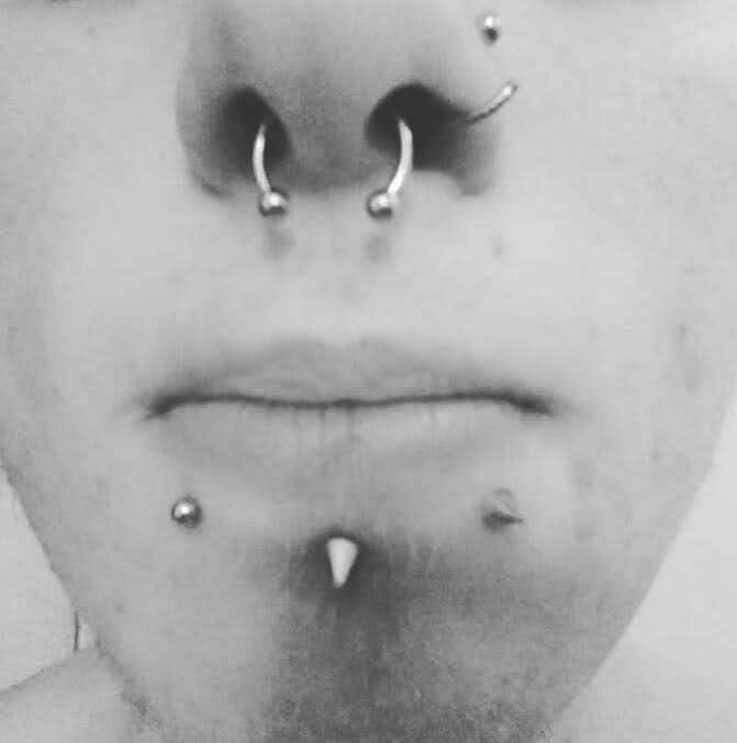 Czarno-białe zbliżenie twarzy z licznymi piercingami: kolczyk w przegrodzie nosowej, dwa kolczyki w nozdrzach, kolczyk w górnej wardze i dwa kolczyki w dolnej części brody.