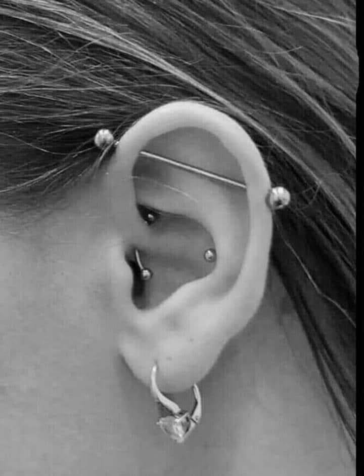 Czarno-białe zbliżenie ucha z licznymi kolczykami, w tym industrial, helix i tragus, oraz wiszący kolczyk w płatku ucha z dużym, błyszczącym kamieniem.