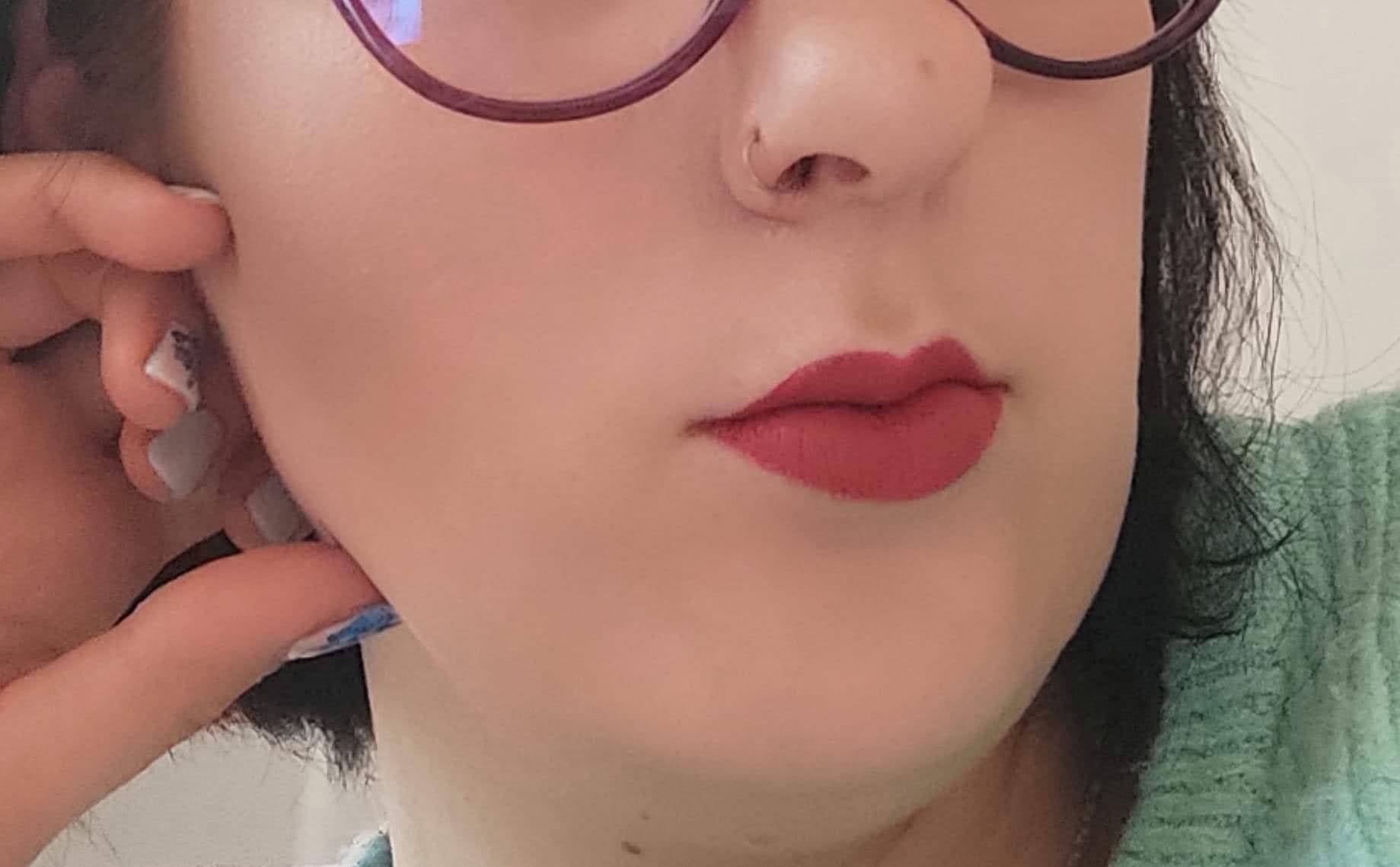 Fragment twarzy kobiety z wyraźnie pomalowanymi na czerwono ustami, kolczyk w nosie i okulary w bordowych oprawkach; paznokcie z delikatnym zdobieniem przy uchu.