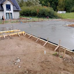 Żelb3t - Bud JAROSŁAW MARCINOWSKI - Wylany fundament pod budowę domu jednorodzinnego, widoczne szalunki, zbrojenie i folia izolacyjna, w tle dom z panelami słonecznymi na dachu.