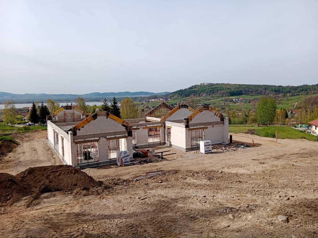 Wznoszenie ścian parterowych domów z bloczków na rozległym, lekko pofałdowanym terenie z widokiem na jezioro i wzgórza w oddali.
