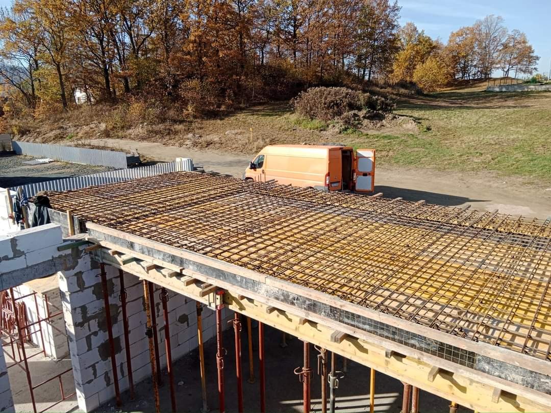 Wiązanie zbrojenia na szalunku stropu budynku, w tle pomarańczowy bus z otwartymi drzwiami i jesienny krajobraz.