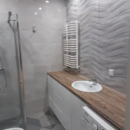 Wykończona łazienka w odcieniach szarości z umywalką nablatową na drewnianym blacie, pod którym znajduje się pralka; na ścianie grzejnik drabinkowy i płytki z falistym wzorem.