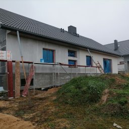 P.k.budownictwo Paweł Kuczyński - Ocieplanie elewacji domu jednorodzinnego w trakcie prac, widoczne rusztowania, zabezpieczone okna folią i częściowo otynkowana ściana szara.