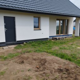 P.k.budownictwo Paweł Kuczyński - Nowoczesny dom jednorodzinny z białą elewacją, ciemną stolarką okienną i grafitowym dachem, widoczna izolacja przeciwwilgociowa fundamentów i częściowo zagospodarowany teren wokół budynku.