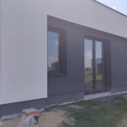 Fragment elewacji domu jednorodzinnego z dwoma oknami, pomalowanego na dwa kolory: biały i grafitowy, z widocznym klimatyzatorem i niedokończonym terenem wokół budynku.