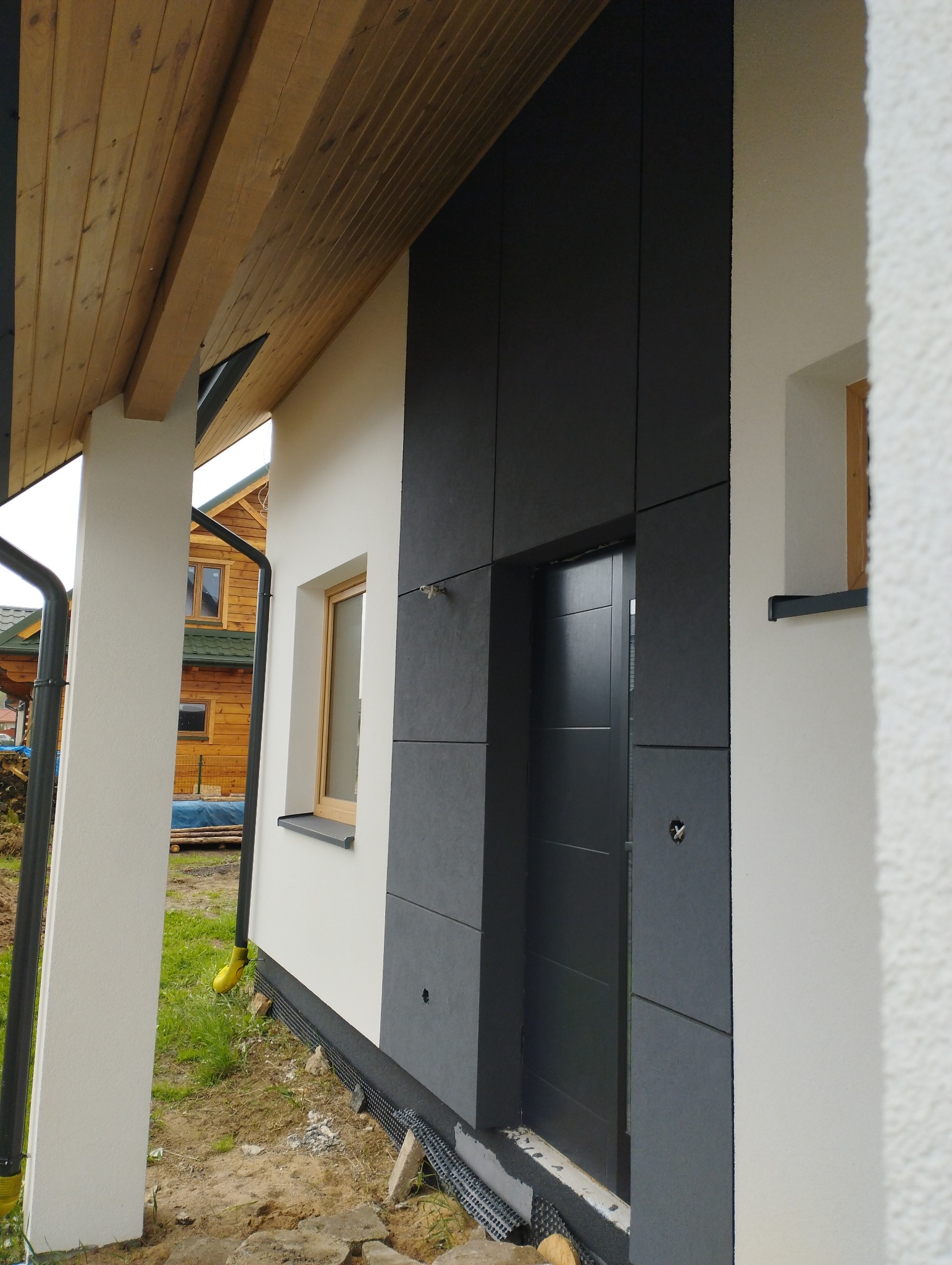 Fragment elewacji domu z szarymi panelami elewacyjnymi i białym tynkiem, widoczna drewniana podbitka i okno z drewnianą ramą, w tle drewniany dom.