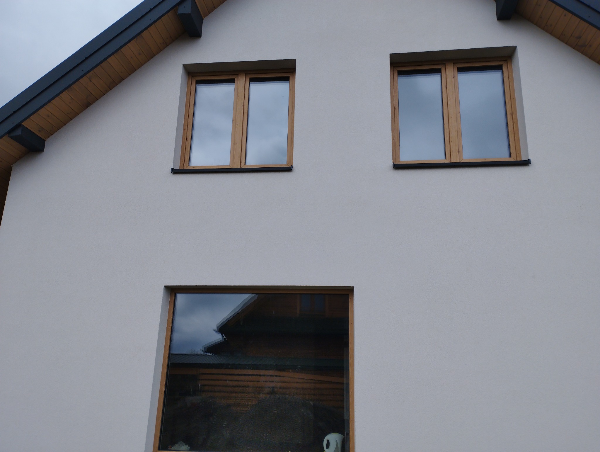 Fragment jasnej elewacji domu z trzema oknami o drewnianych ramach; w dolnym oknie odbija się sąsiedni budynek.