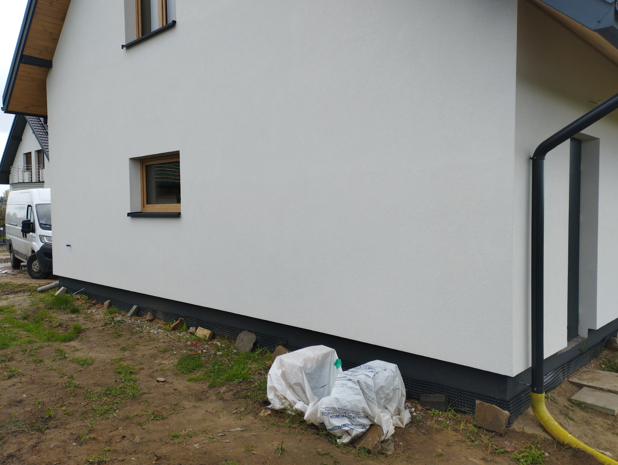 Boczny widok domu z białą elewacją i drewnianymi oknami, widoczne zabezpieczone materiały budowlane przy fundamencie oraz zaparkowany biały bus.