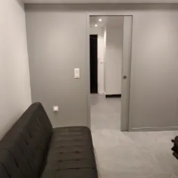 Szare wnętrze mieszkania z sofą i przesuwnymi drzwiami do kolejnego pomieszczenia, widoczne gładkie ściany i podłoga z płytek.