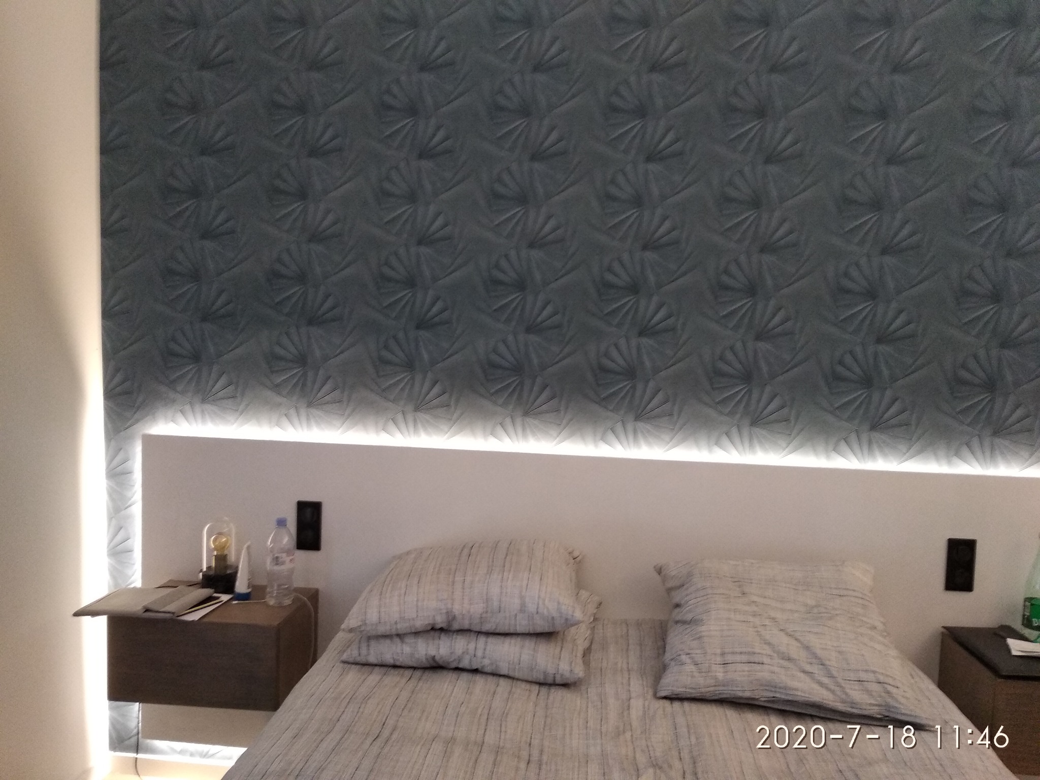 Sypialnia z szarą tapetą o geometrycznym wzorze, podświetlana listwą LED nad wezgłowiem łóżka, z widocznymi stolikami nocnymi i elementami dekoracyjnymi.