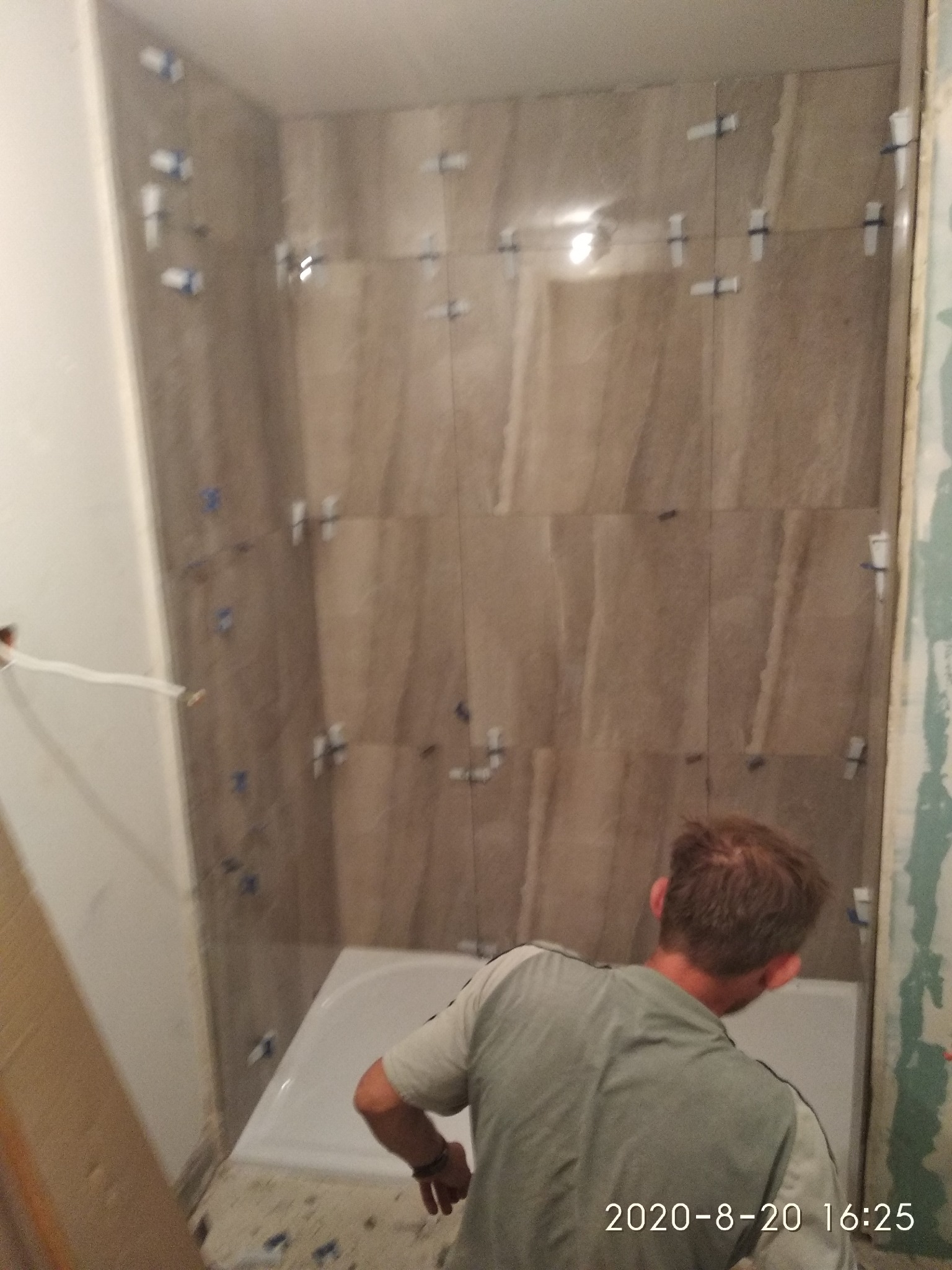 Montaż płytek imitujących kamień na ścianie w łazience, widoczne plastikowe krzyżyki dystansowe, biała wanna i fragment sylwetki instalatora.