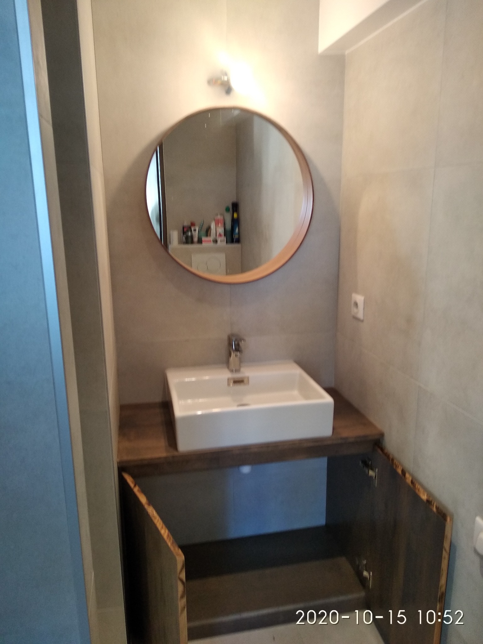 Nowoczesna łazienka z kwadratową umywalką nablatową, okrągłym lustrem w drewnianej ramie i szafką z otwartymi drzwiczkami ukazującą półkę wewnątrz.