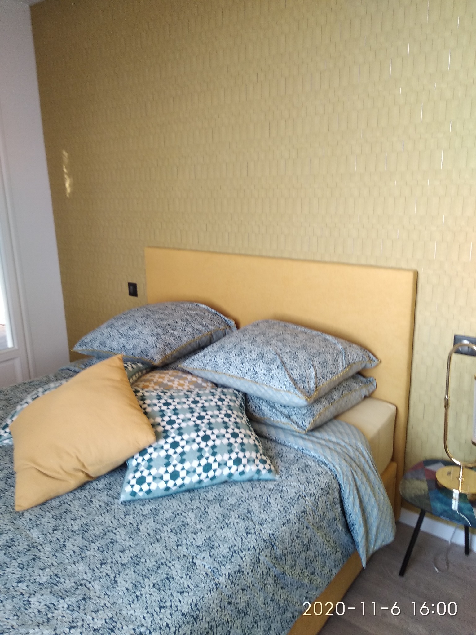 Sypialnia z łóżkiem z żółtym zagłówkiem, wzorzystą pościelą i złotą tapetą w pionowe prążki.