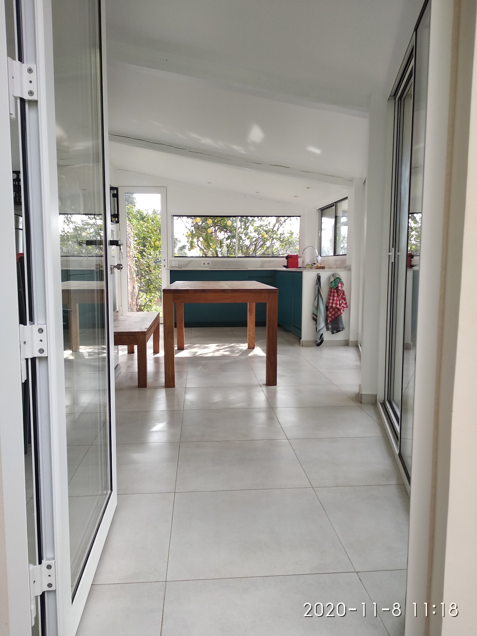 Jasne wnętrze z otwartymi, składanymi drzwiami, ukazujące minimalistyczną kuchnię z drewnianym stołem i niebieskimi szafkami, widok na ogród przez okno.