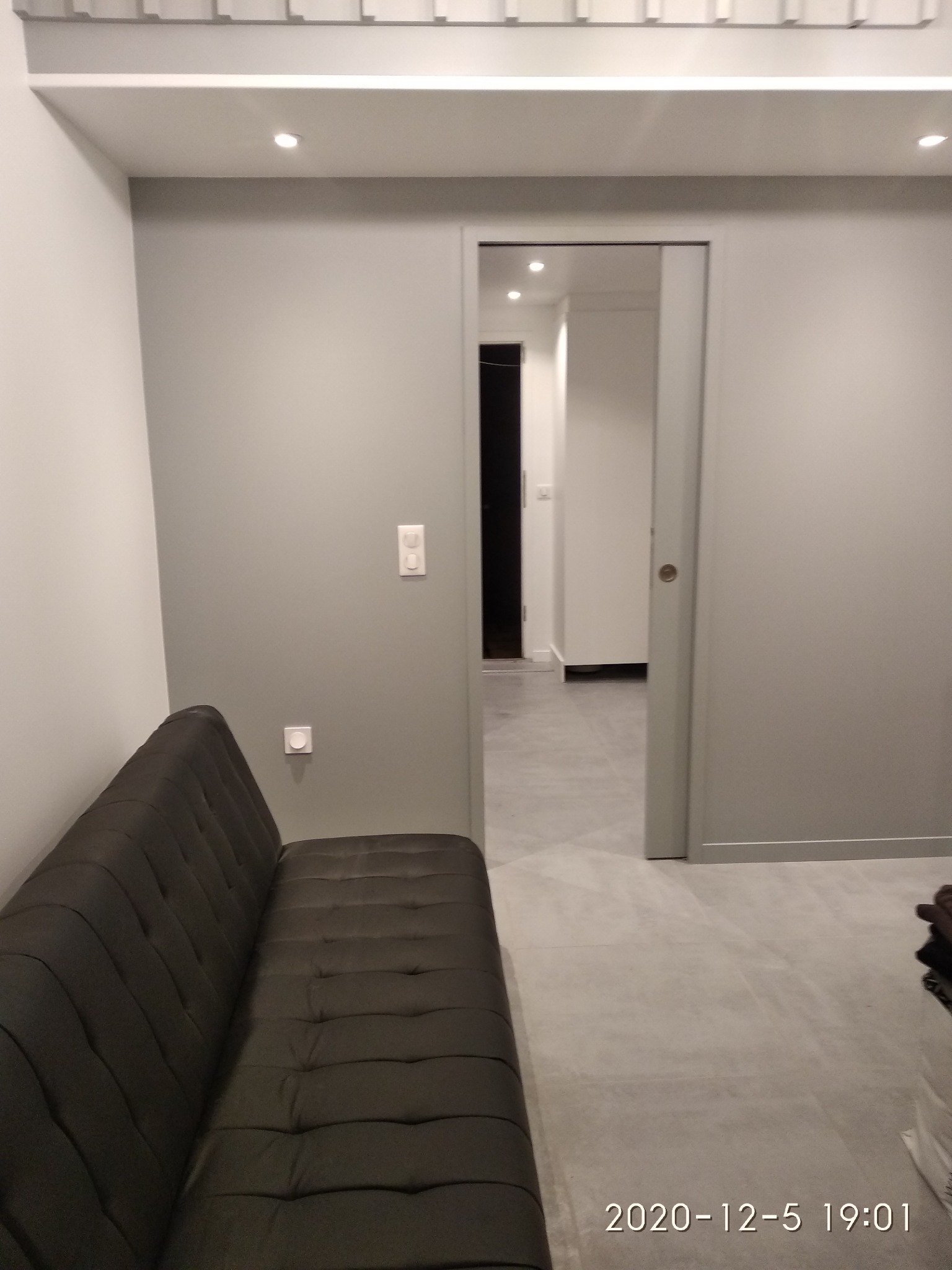 Szare wnętrze mieszkania z sofą i przesuwnymi drzwiami do kolejnego pomieszczenia, widoczne gładkie ściany i podłoga z płytek.