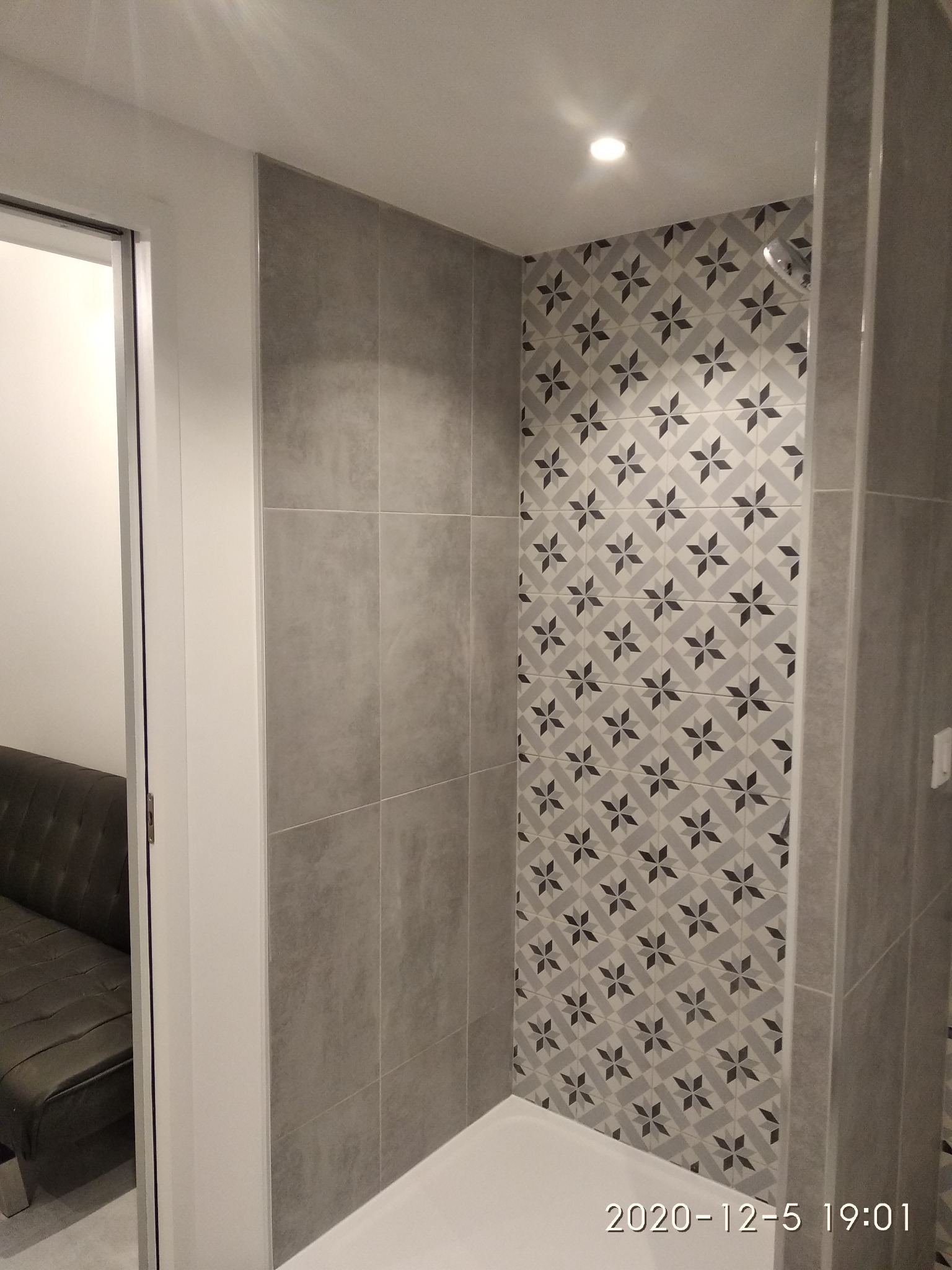 Wykończona kabina prysznicowa z szarymi płytkami o wzorze geometrycznym, oświetlona punktowym światłem, widok z boku, fragment sofy widoczny w tle.