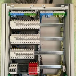 Otwarta skrzynka elektryczna z zainstalowanymi bezpiecznikami, różnicówkami i przewodami w kolorach niebieskim, żółtym i zielonym, zamontowana na ścianie z widocznymi rurami karbowanymi