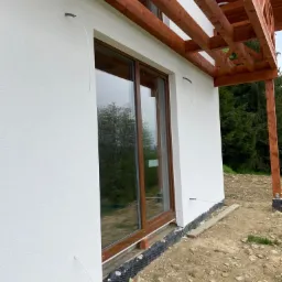 Biała elewacja budynku z widocznymi przewodami wychodzącymi ze ściany, prawdopodobnie przygotowanymi do podłączenia zewnętrznego oświetlenia lub innych urządzeń. Widoczne drewniane elementy...