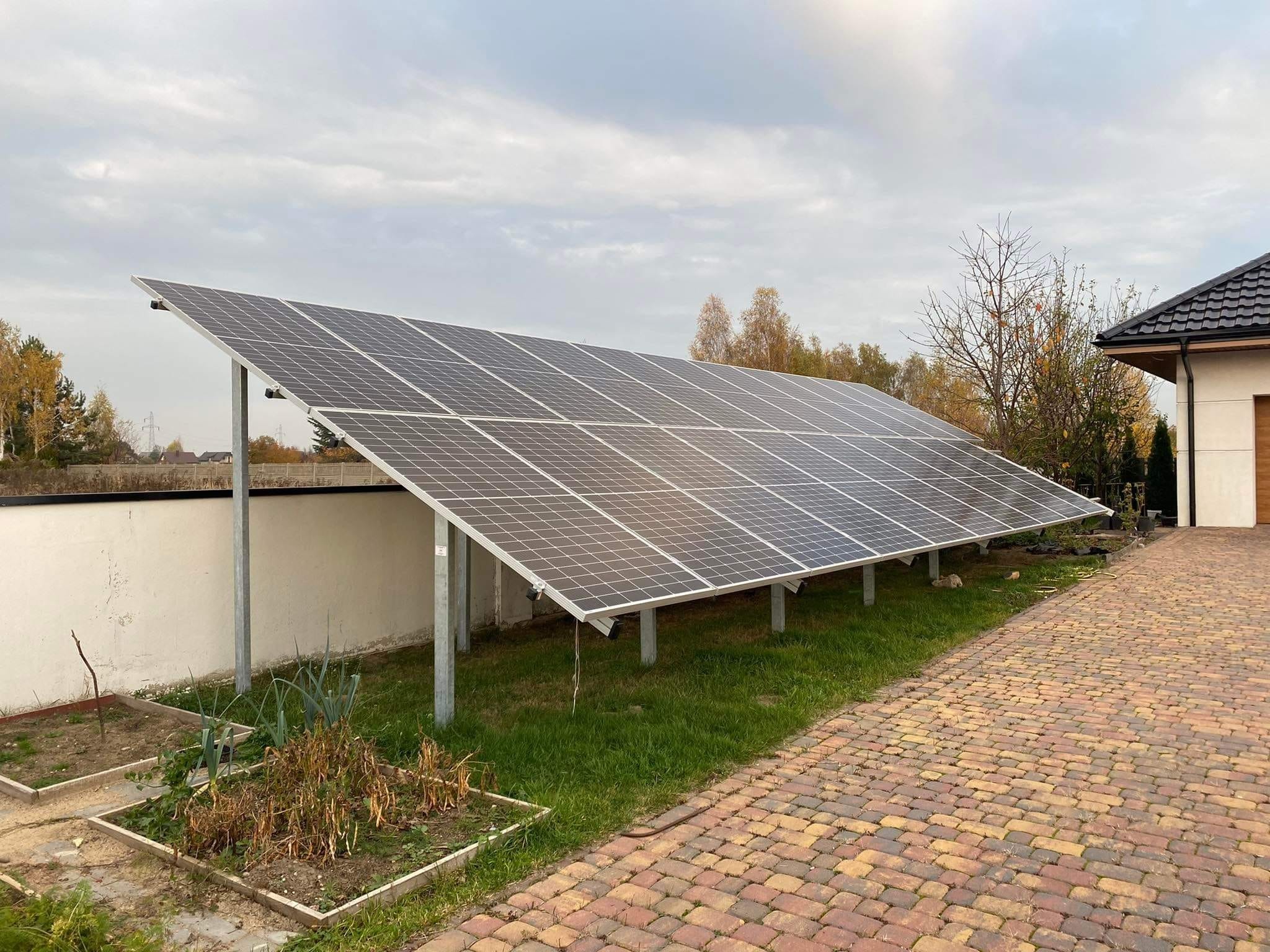 Rząd paneli słonecznych zamontowanych na metalowych wspornikach, ustawionych na trawniku obok domu z brukowanym podjazdem i małym ogródkiem warzywnym z przodu.