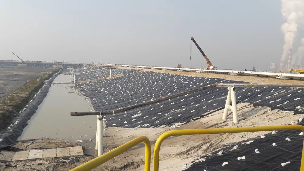 Budowa lub rekultywacja składowiska odpadów: czarna folia zabezpieczająca z białymi obciążnikami, rury odprowadzające, zbiornik wodny, w tle kominy i dźwig budowlany.