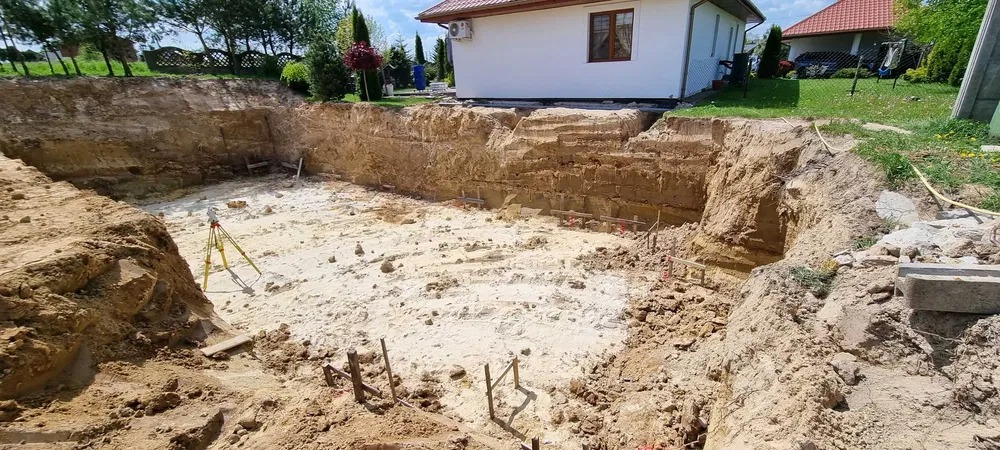 Wykop pod fundamenty domu jednorodzinnego z widocznym niwelatorem optycznym na trójnogu, drewnianymi rozpórkami i odsłoniętymi warstwami gruntu.