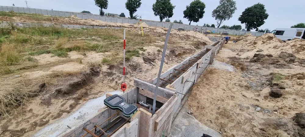 Wykonanie fundamentów z deskowaniem i zbrojeniem, widoczny kontroler pomiarowy i łata geodezyjna, w tle teren budowy z częściowo wzniesionym murem.