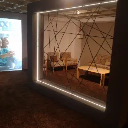 Aranżacja wnętrza z dekoracyjną ścianką działową podświetloną LED, widoczna część ekspozycji mebli tapicerowanych i reklama świetlna w tle.