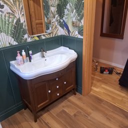 FESTBUD JAROSŁAW RYBICKI - Umywalka z szafką w łazience z egzotyczną tapetą w liście i zwierzęta, drewniana podłoga, widoczna skrzynka z narzędziami w przejściu do innego pomieszczenia.