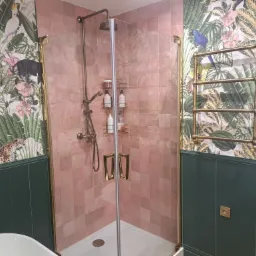 Nowoczesna łazienka z kabiną prysznicową wykończoną różowymi płytkami, zielonymi panelami ściennymi i tapetą w motywy tropikalne. Widoczny grzejnik drabinkowy w kolorze starego złota i bateria...