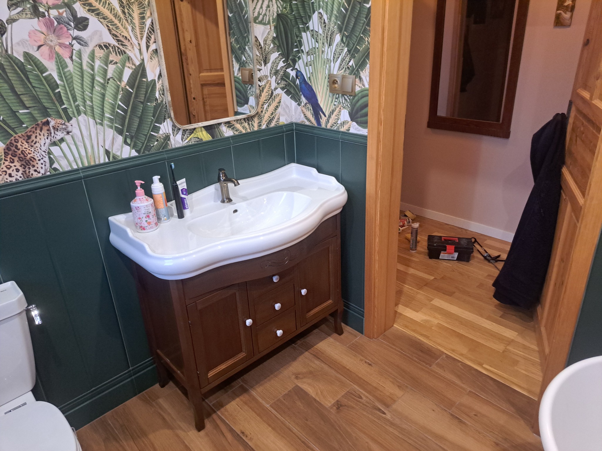 Umywalka z szafką w łazience z egzotyczną tapetą w liście i zwierzęta, drewniana podłoga, widoczna skrzynka z narzędziami w przejściu do innego pomieszczenia.