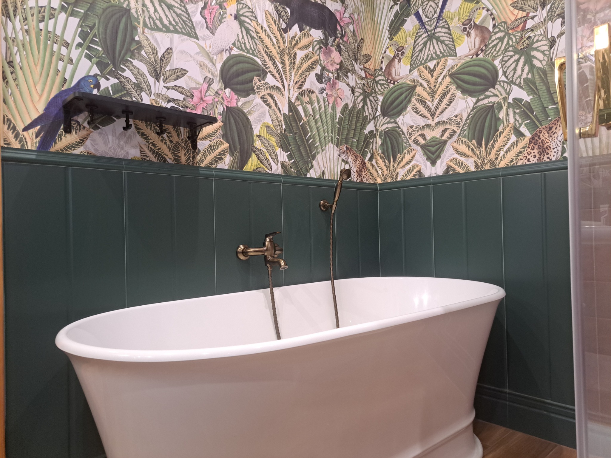 Wolnostojąca wanna w łazience z ciemnozielonymi płytkami i tapetą w tropikalne wzory z motywami zwierzęcymi, armatura w kolorze starego złota.