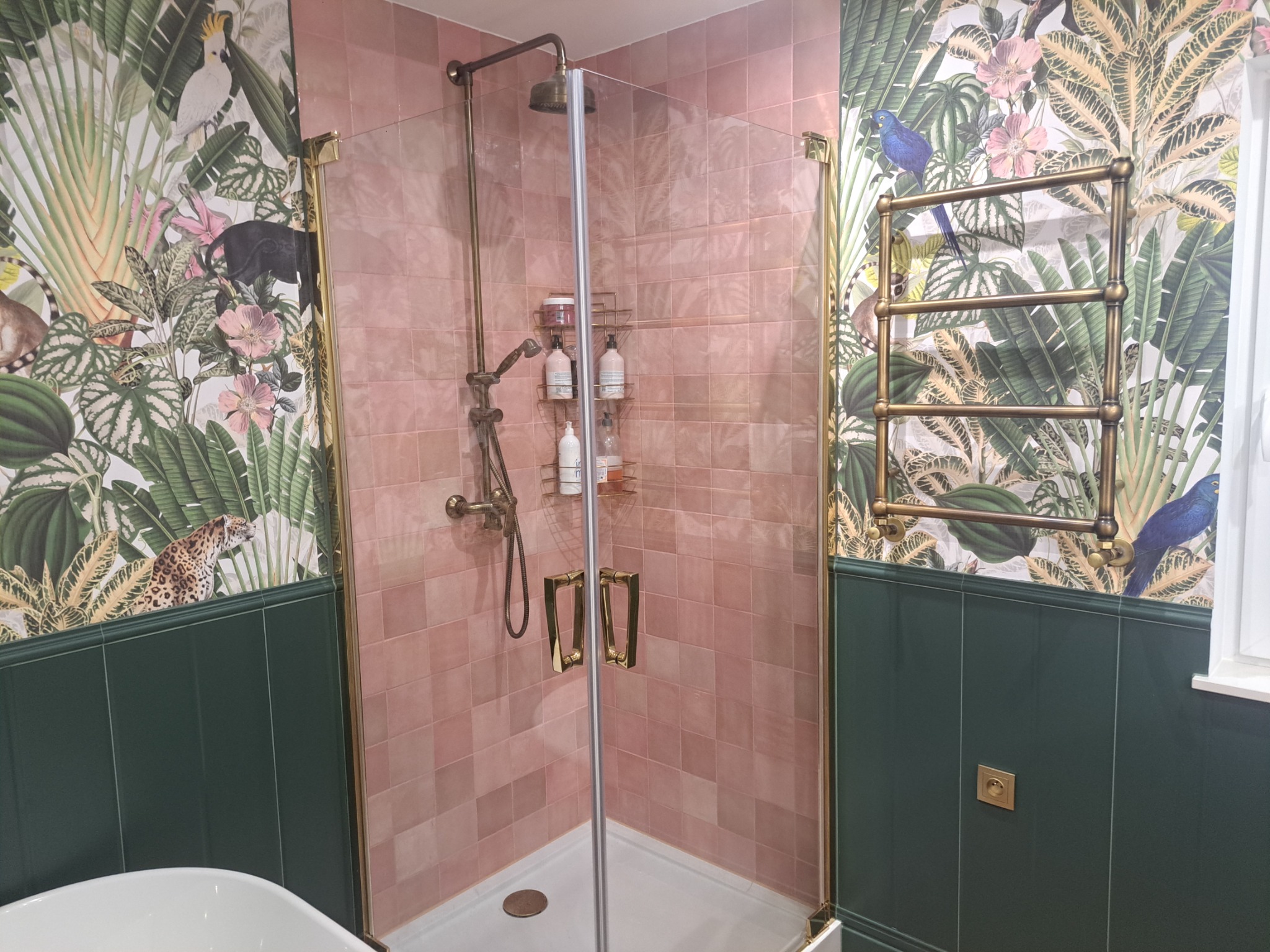 Nowoczesna łazienka z kabiną prysznicową wykończoną różowymi płytkami, zielonymi panelami ściennymi i tapetą w motywy tropikalne. Widoczny grzejnik drabinkowy w kolorze starego złota i bateria...