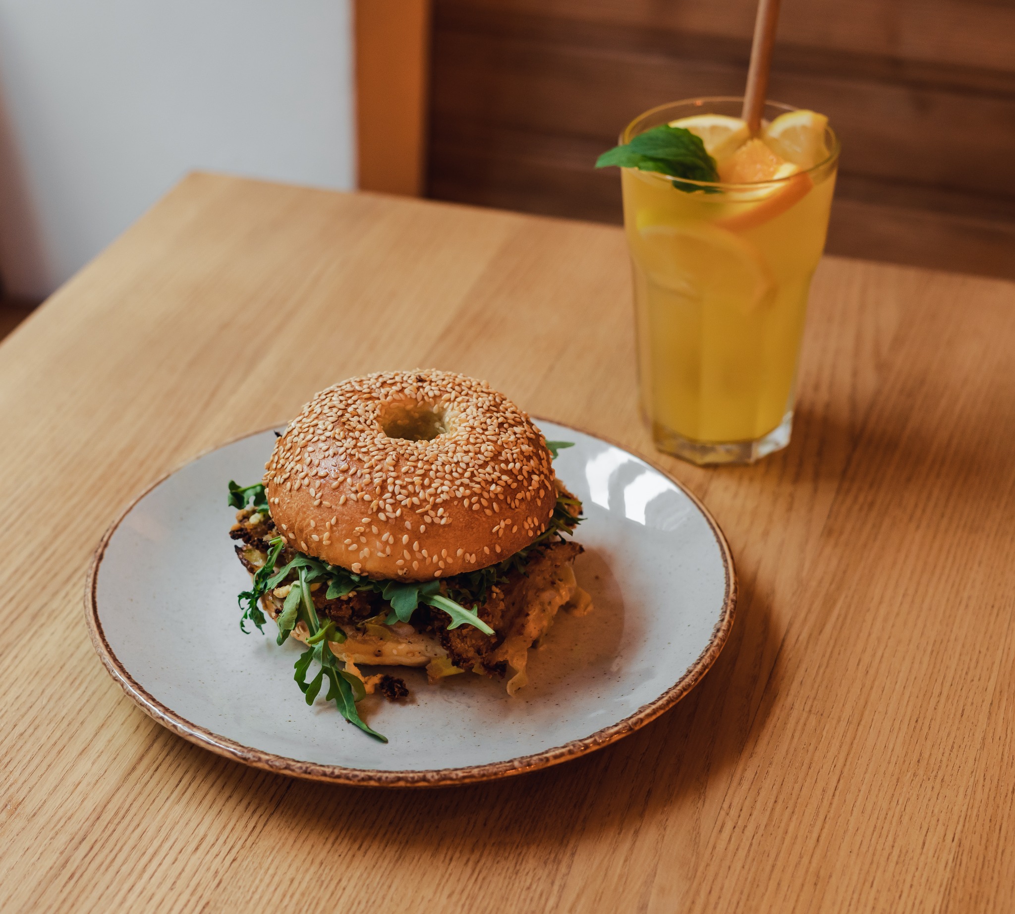 Bagel z sezamem wypełniony warzywami i sosem na talerzu obok szklanki lemoniady z pomarańczą, cytryną i miętą, całość na drewnianym stole.