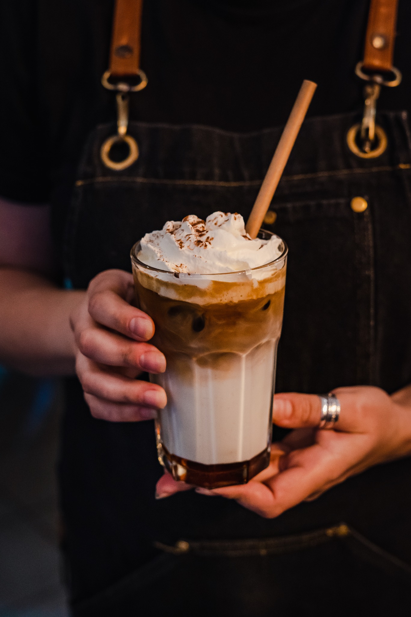 Barista w dżinsowym fartuchu prezentuje szklankę mrożonej kawy z bitą śmietaną i cynamonem, podtrzymując ją dłońmi z pierścionkiem.